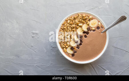 Schokolade smoothie Schüssel mit Bananen, Müsli und Erdnüsse auf Hellgrau konkreten Hintergrund. Gesundes vegetarisches Frühstück. Ansicht von oben mit Platz für Tex Stockfoto