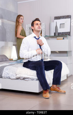 Mann sitzt auf dem Bett und putzt sich, während seine Freundin prinking vor Spiegel Stockfoto