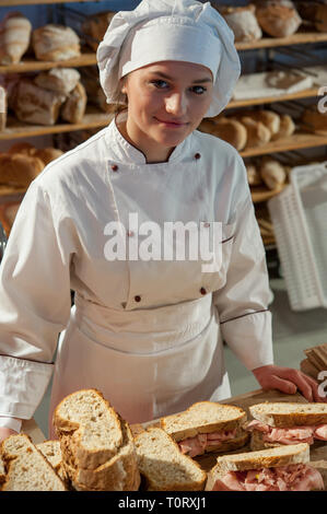 Lächelnden jungen Verkäuferin in der Bäckerei Schreibtisch bereitet Mortadella Bologna Sandwiches. Regale voll Brot in den Hintergrund. Stockfoto