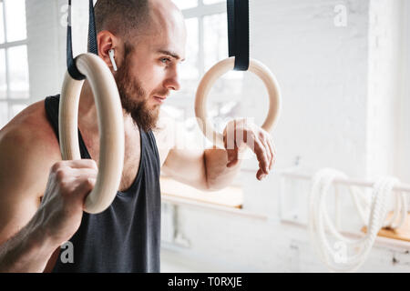Müde passen Mann hält gymnast Ringe und weg schauen. Bärtige Athleten mit drahtlosen Kopfhörern trainieren im Fitnessstudio Stockfoto