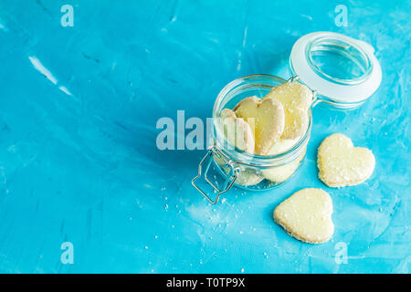 Köstliche gebackene Kekse in Herzform mit Kokos Chips in Glas Glas auf blauen Beton Tabelle Oberfläche. Essen Grußkarte mit kopieren Sie Text. Stockfoto