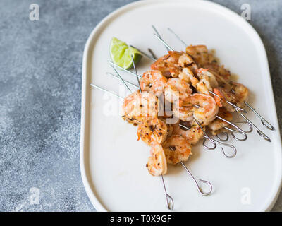 Gegrillte pikante Garnelen auf Metall Spieße mit Petersilie und Limettensaft auf ovalen weißen Teller auf grauem Hintergrund. Stockfoto