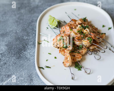 Gegrillte pikante Garnelen auf Metall Spieße mit Petersilie und Limettensaft auf ovalen weißen Teller auf grauem Hintergrund. Stockfoto