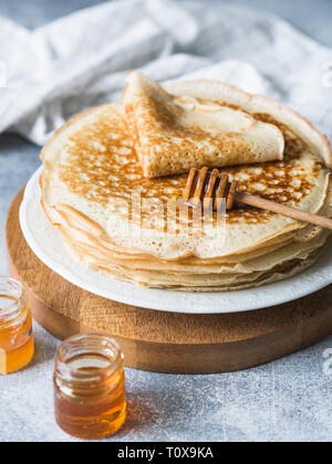 Stapel Russischen hausgemachter Hefe Pfannkuchen auf weiße Platte, Holz Honig Löffel und Honig in Gläsern auf dem Tisch. Traditionellen Weizen Pfannkuchen für Fastnacht. Stockfoto