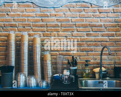 Reihen von Stapel von oben nach unten aus transparentem Kunststoff Kaffee Tasse in der Nähe von Edelstahl Spüle auf Mauer Hintergrund im Cafe mit kopieren. Stockfoto