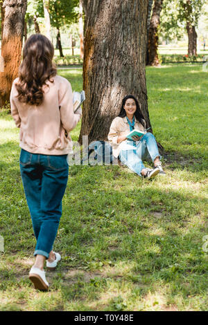 Rückansicht der Mädchen gehen mit Büchern in der Nähe von fröhlichen Freund lächelnd, während er sich in der Park sitzen Stockfoto