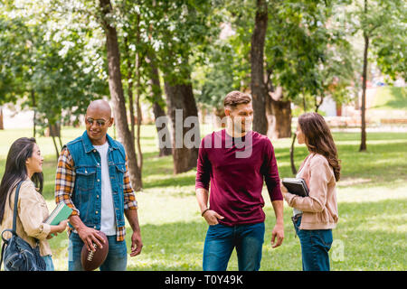 Attraktive Mädchen reden mit fröhlichen multikulturellen Studenten beim Stehen mit Büchern in Park Stockfoto