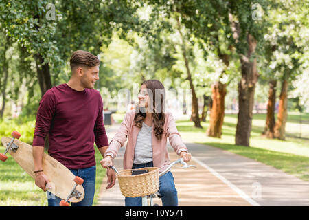 Schöner Mann holding Longboard und Suchen zu attraktiven Freundin Reiten Fahrrad Stockfoto