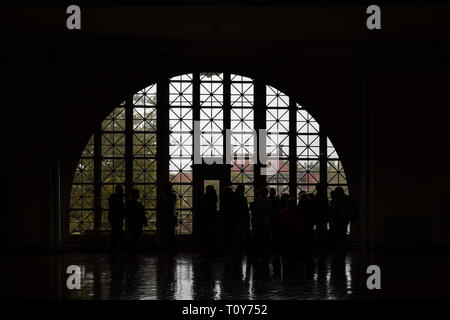 Touristen in Silhouette aus einem Halbkreis kunstvoll verglasten Fenster in der Registry Zimmer von Ellis Island in New York City. Stockfoto