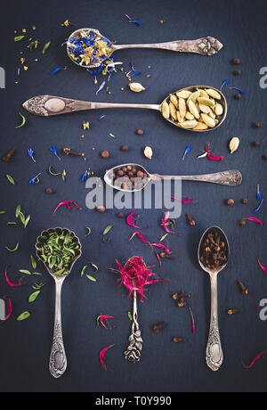 Silber Löffel voller Gewürze, Kräuter und Tees, Blumen auf schwarzem Schiefer Hintergrund. Stockfoto