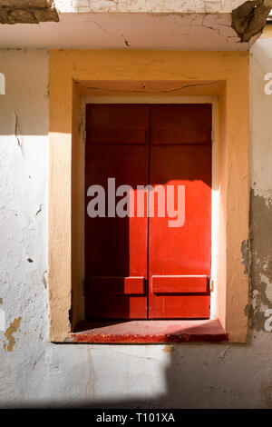 Wand von einem älteren Haus mit einem Fenster von einem hellen Rot Holz- Blende abgedeckt. Orange Rahmen. Starker Sonneneinstrahlung. Straße in Rethymnon, Kreta, Griechenland. Stockfoto