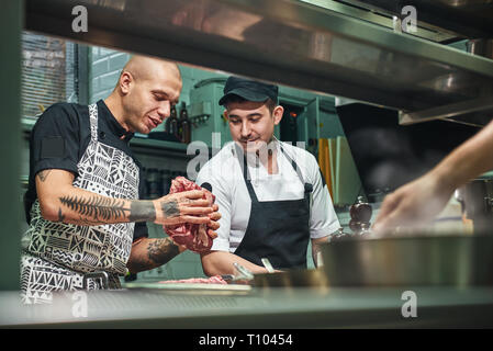Wie Sie das richtige Fleisch professioneller Koch in Schürze und mit Tattoos auf seine Arme ein rotes Fleisch zu seinem Assistenten wählen Sie beim Stehen in einem Restaurant Küche. Stockfoto