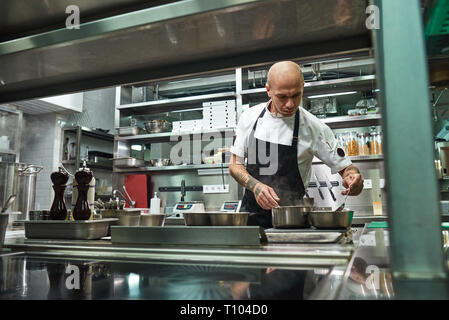 Restaurant Chef. Konzentrierten jungen Koch in schwarze Schürze ist Kochen in seinem modernen Restaurant Küche. Kochen Konzept Stockfoto