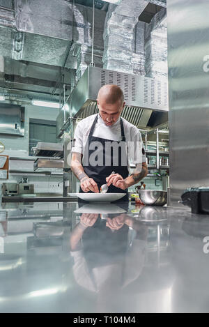 Die Arbeit in einem Restaurant. Vertikale Portrait von professionellen männlichen Chef mit Tattoos auf seinem Arme garnieren seinen Teller auf die weiße Platte, während in der Küche stehend Stockfoto