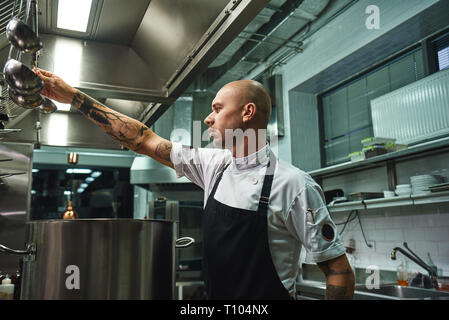 Berühmte Suppe. Seitenansicht des attraktiven kahlen Koch Schürze, einen Löffel zum Kochen von Suppe beim Stehen in einem Restaurant Küche. Kochen Konzept Stockfoto