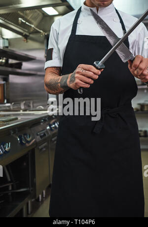 Bereit zu kochen. Vertikale Foto des jungen Mannes in Schürze mit Tattoos auf seinem Arme Schärfen eines Messers in einem Restaurant Küche. Kochen Konzept Stockfoto