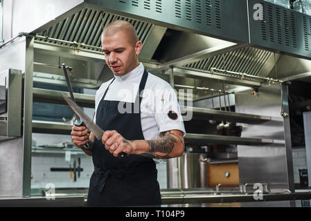 Alles sollte perfekt Brutale Chef mit mehreren Tattoos auf seinem Arme Schärfen eines Messers während in einem Restaurant in der Küche stehen. Kochen Konzept Stockfoto