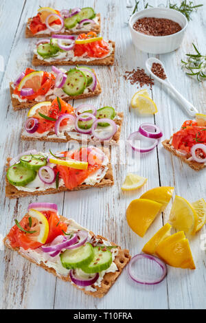 Knäckebrot Open-faced Sandwich mit geräucherter Forelle, weiche Creme Käse, Gurken, rote Zwiebel, bestreut mit Leinsamen auf einem weißen alten Holztisch, Verti Stockfoto