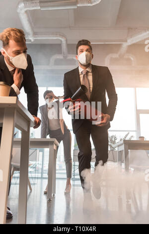 Geschäftsmann in Maske holding Feuerlöscher in der Nähe von Kollegen im Büro mit Rauch Stockfoto