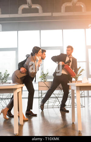 Erschrocken Geschäftsmann holding Feuerlöscher in der Nähe von Kollegen beim Gehen im Büro mit Rauch Stockfoto