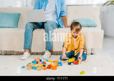 Vater saß auf der Couch und mit Laptop während Vorschüler Sohn spielen mit bunten Bausteinen zu Hause Stockfoto