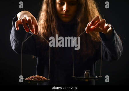 Emotionale Portrait eines Mädchens mit langen Locken mit einem Gewicht von Korn und Gewichte für Waagen vor einem schwarzen Hintergrund, Low Key Stockfoto