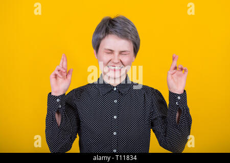 Gerne attraktive junge Frau den Wunsch zu gewinnen, hebt die Hände mit gekreuzten Fingern, wartet auf Lotterie Ergebnisse, schließt die Augen, kurze Frisur, gekleidet Stockfoto