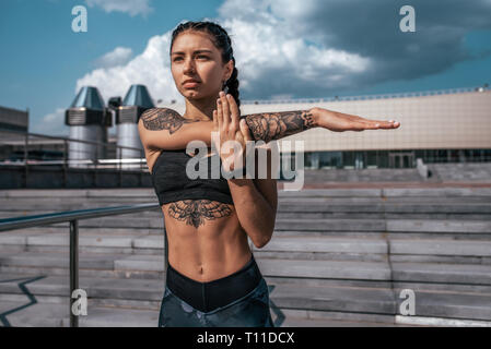 Schöne Mädchen, Athlet, gebräunte Haut Tattoos. Es wärmt die Muskeln vor dem Training im Sommer in der Stadt. Ellbogen und Schulter, Gelenke, Gelenkentzündungen Gymnastik. Sicher Stockfoto