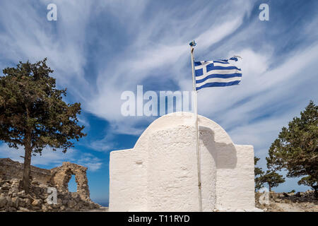 Die Apsis einer typischen griechischen Kirche mit der Fahne im Wind unter den Ruinen der mittelalterlichen Burg Monolithos, gegen eine dramatische Himmel, Insel Rhodos, Stockfoto