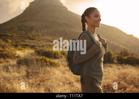 Eine Frau mit einem Rucksack auf den Schultern hinunter Landstraße in der Mitte eines Feldes mit Gras. Auf querfeldein Spaziergang im Sommer Tag weiblich. Stockfoto