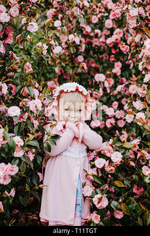 Hübsches kleines Mädchen mit rosa Motorhaube und rosa Kleid in der Blüte Bush Rosa Camelia in sonniger Frühlingstag Stockfoto