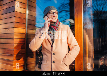 Junge stilvolle Geschäftsmann mit Smartphone und Berufung durch die Cafe. Hübscher Kerl trägt klassische Kleidung und Accessoires im Freien Stockfoto