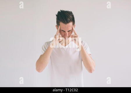 Mann mit Brille Probleme und Kopfschmerzen zu Hause. Junge schwarzhaarige nerd Kerl tragen weiße t-shirt, Hände auf den Kopf, während sich über s gefunden Stockfoto