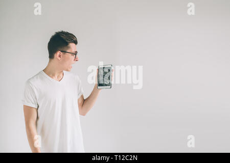 Nerd eine Brille trägt. Schüler, die eine gebrochene schwarz Tablet hinter Glas. umgekippt Mann hält eine Out-of-use Tablet-PC oder Smartphone. Auf einem hellen Isoliert Stockfoto