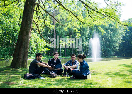 Eine Gruppe von jungen, internationalen Studenten sitzen draußen auf College Campus unter einem schattigen Baum auf dem Gras Stockfoto