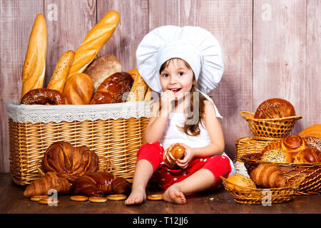 Brot rund um den Kopf. Wenig lächelnde Mädchen in einer Cook cap isst Brot und Bagels in der Nähe von einem Weidenkorb mit Brötchen und Backwaren. Stockfoto