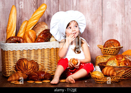 Brot rund um den Kopf. Wenig lächelnde Mädchen in einer Cook cap isst Brot und Bagels in der Nähe von einem Weidenkorb mit Brötchen und Backwaren. Stockfoto