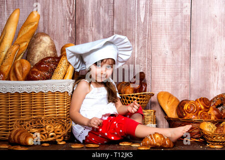 Brot rund um den Kopf. Wenig lächelnde Mädchen in einer Cook cap isst Brot und Bagels in der Nähe von einem Weidenkorb mit Brötchen und Backwaren. Stockfoto