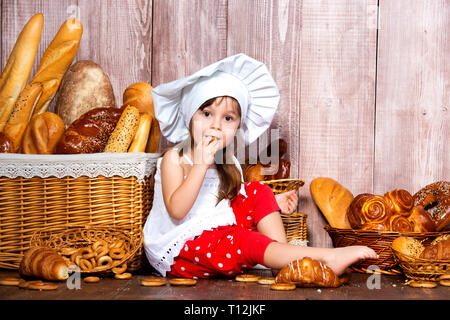 Brot rund um den Kopf. Wenig lächelnde Mädchen in einer Cook cap isst Brot und Bagels in der Nähe von einem Weidenkorb mit Brötchen und Backwaren. Stockfoto