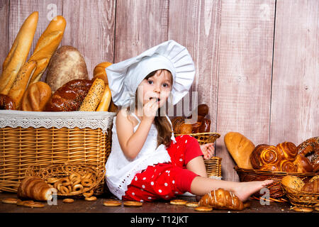 Brot rund um den Kopf. Wenig lächelnde Mädchen in einer Cook cap isst Brot und Bagels in der Nähe von einem Weidenkorb mit Brötchen und Backwaren. Stockfoto