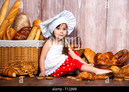 Brot rund um den Kopf. Wenig lächelnde Mädchen in einer Cook cap isst Brot und Bagels in der Nähe von einem Weidenkorb mit Brötchen und Backwaren. Stockfoto