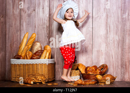 Ich liebe Brötchen. Wenig lächelnde Mädchen in einem kochenden Gap springen für Freude und Vergnügen in der Nähe von einem Weidenkorb mit Brötchen und Backwaren. Stockfoto