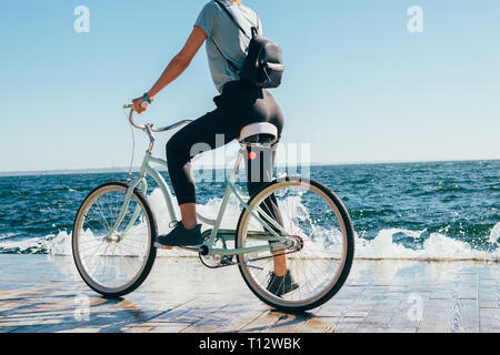 Rückansicht junge Frau während der Fahrt mit dem Fahrrad an der sonnigen Sommertag gestoppt. Frauen tragen Rucksack stehend einen Fuß auf Pedal von Cruiser Fahrrad in der Nähe von Blue se Stockfoto