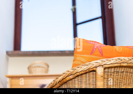 Eine komfortable vintage Stroh Sessel home oder Haus Wohnzimmer oder Schlafzimmer durch offene Fenster und Himmel mit Strand orange Kissen closeup Stockfoto