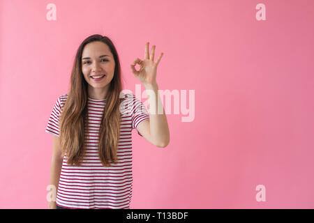 Positive lächelnde Mädchen auf einem rosa Hintergrund zeigt Finger Geste Bedeutung Ok. Stockfoto