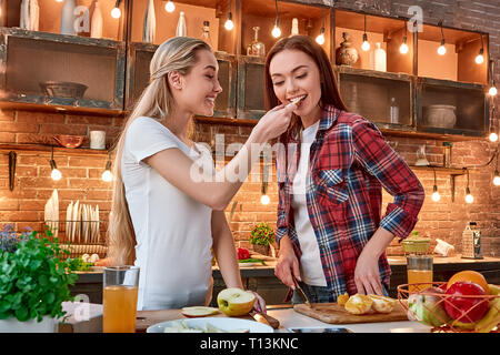 Porträt von zwei attraktive Frauen, Spaß zu haben, während der Vorbereitung Obstsalat. Sie sind voll in den Prozess einbezogen. Blonde Mädchen im weißen T-Shirt ist Ihr dunkelhaarigen Freund in kariertem Hemd, mit Apfel Schicht, während sie Verkostung ist es. Ansicht von vorn Stockfoto