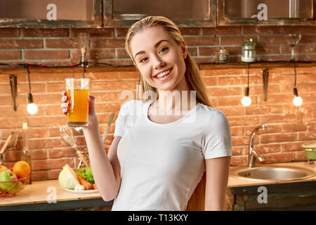 Portrait von fröhlichen blonde Mädchen tragen weiße T-Shirt, mit frischem Saft, mit Blick auf die Kamera und lächelnd. Attraktive junge Frau bereitet sich auf gesundes Essen. Grün gesund essen Konzept. Ansicht von vorn Stockfoto