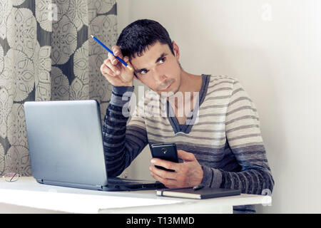 Portrait des jungen Mannes in Freizeitkleidung auf dem Job Arbeiten am Laptop, Smartphone und mit nachdenklichen Gesicht Stockfoto