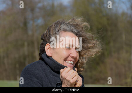 Lächelnde Frau mittleren Alters an einem windigen Tag auf dem Lande, kuschelte sich in eine warme Jacke, wie ihr Haar weht im Wind frei Stockfoto