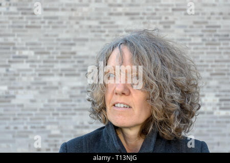 Reifen serios Frau mit Curly zerzaustem Haar auf die Seite, wie Sie sich vor einer grauen Wand mit Kopie Raum Posen Stockfoto